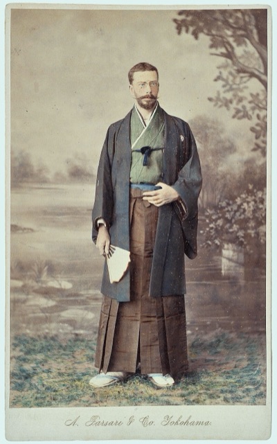 Hand-coloured studio photograph of a bearded European man in spectacles, wearing a kimono and hakama and holding a fan.