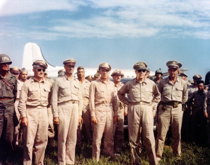 Colour photograph of General Douglas MacArthur in khaki uniform and sunglasses, standing with other American officers on an airfield, an aircraft behind them.