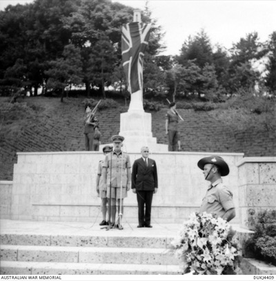 Black-and-white photograph of Lieutenant General Sir Horace Robertson in uniform at a microphone in front of a memorial cross, with a guard of honour and a Union flag.